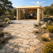 Rectangular light wood studio with glass sliding doors and a flat roof, set on a stone patio in a garden landscape.