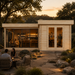 Rectangular light wood cabin with glass doors and windows at sunset, featuring people inside and a stone patio outside.