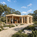 L-shaped wood cabin with glass walls and doors used as an office, set in a landscape with trees and a gravel path.