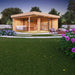 A cozy, modern GardenHouse24 USA ALU Concept Relax 70A Garden Room with large glass doors, a hanging chair, indoor office setup, and soft evening, with a grass lawn and lighting surrounded by blooming flowers.