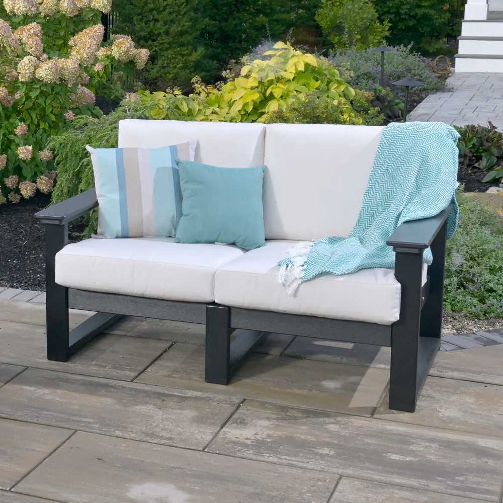 A LuxCraft Lanai Deep Seating Loveseat is placed in a garden with lush greenery, accented with soft blue and striped pillows