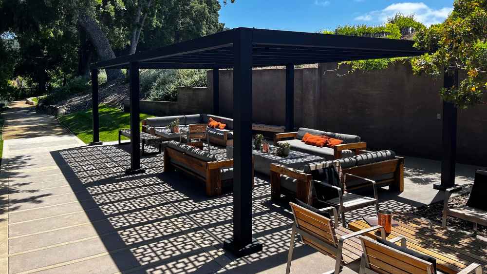 A Structureworks Shadow Trex Pergola casts bold geometric shadows over a cozy outdoor lounge with cushioned wooden seating and a sleek black frame