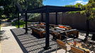 A Structureworks Shadow Trex Pergola casts bold geometric shadows over a cozy outdoor lounge with cushioned wooden seating and a sleek black frame