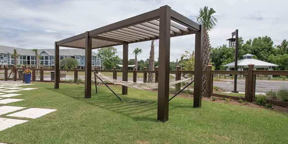 A Structureworks Stratus Aluminum Pergola stands over a hammock in a grassy yard near palm trees and a wooden fence