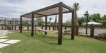 A Structureworks Stratus Aluminum Pergola stands over a hammock in a grassy yard near palm trees and a wooden fence