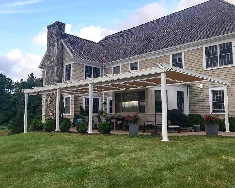 A beige-roofed Structureworks Atlas Aluminum Pergola shades a wide back patio attached to a large, traditional-style house