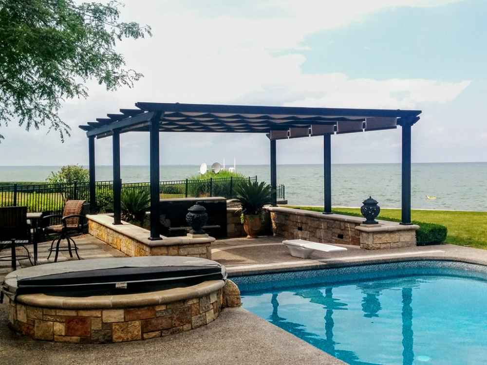 A black Structureworks Atlas Aluminum Pergola with a patterned canopy stands beside a pool, bordered by a stone wall and facing a lake
