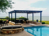 A black Structureworks Atlas Aluminum Pergola with a patterned canopy stands beside a pool, bordered by a stone wall and facing a lake