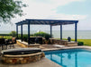 A black Structureworks Atlas Aluminum Pergola with a patterned canopy stands beside a pool, bordered by a stone wall and facing a lake