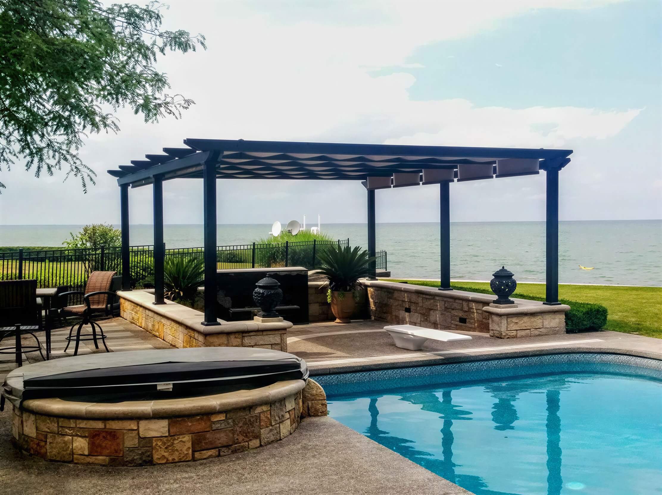 A black Structureworks Atlas Aluminum Pergola with a patterned canopy stands beside a pool, bordered by a stone wall and facing a lake