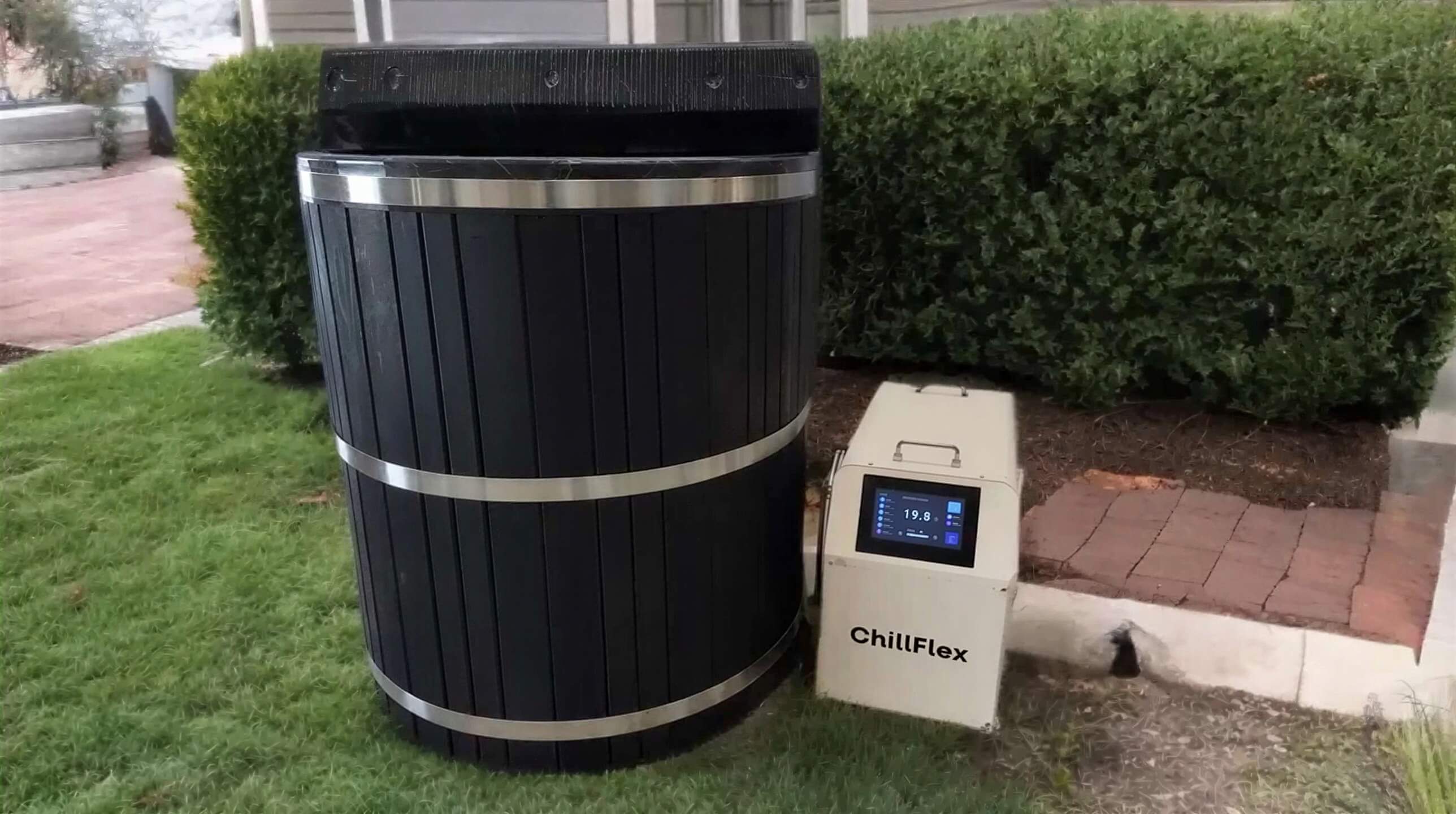 A black barrel-style Sawmill Structures Chiller Plunge with a ChillFlex cooling unit sits neatly on a green lawn near a home