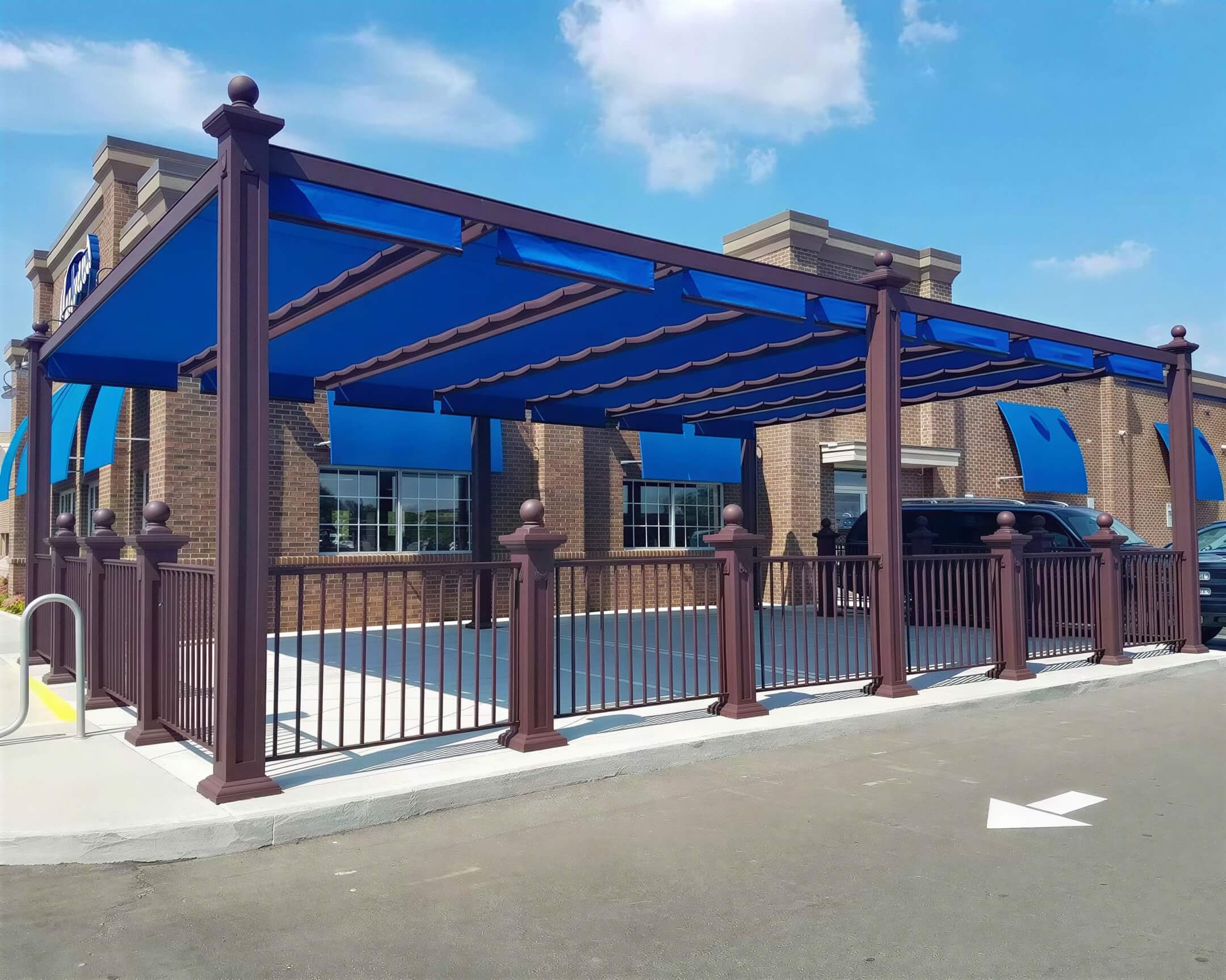 A blue-canopied outdoor seating area in front of a brick building, protected by a Structureworks Windsor Aluminum Patio Cover with matching awnings