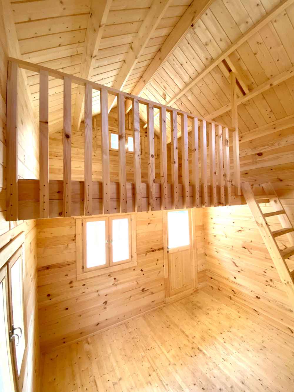 A bright, empty interior with tall walls, a wood ladder, and a railed loft showcases the spacious design of the Sawmill Structures The Jumbo Grand Loft 130 SD Garden Room