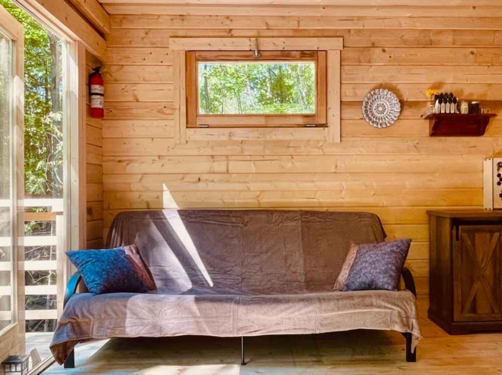 A brown-covered futon with blue and purple pillows sits under a small window inside the Sawmill Structures Edwin Lake House 70MM Garden Room