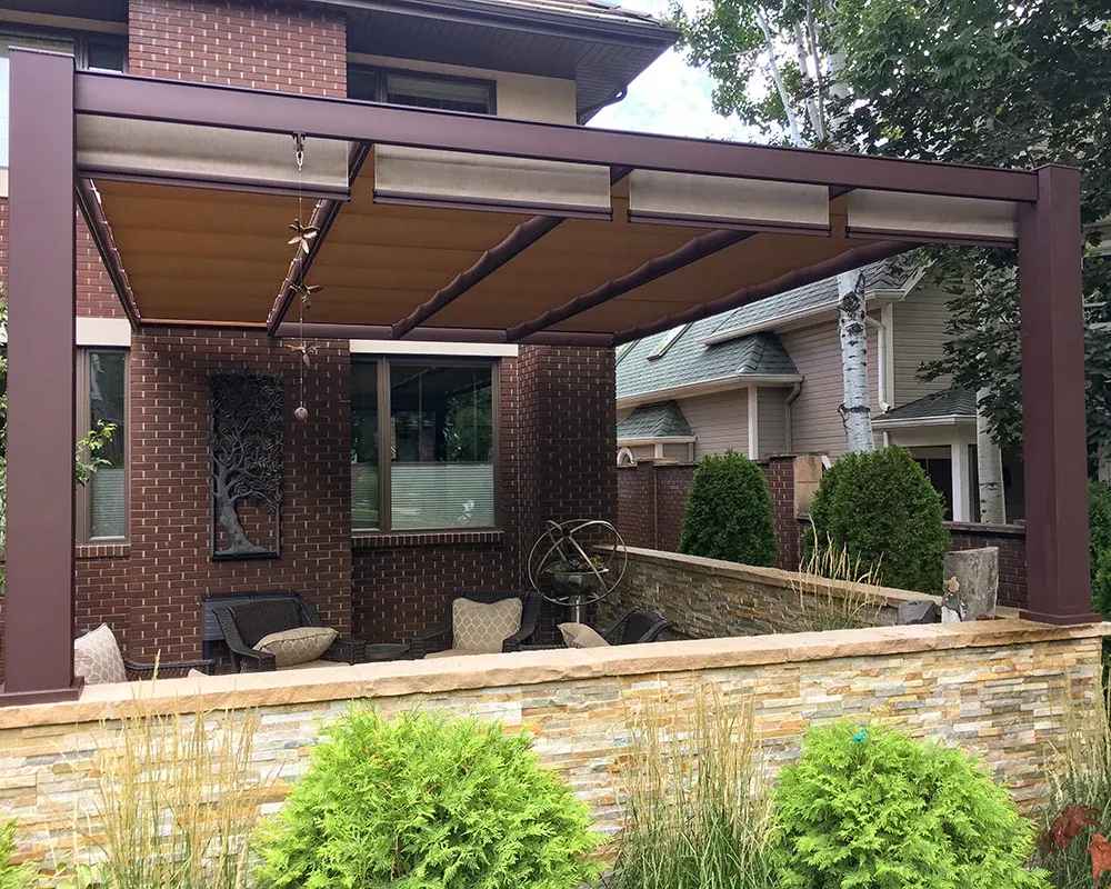 A brown Structureworks Outlook Aluminum Patio Cover & Pergola covers a cozy sitting area in a private courtyard surrounded by stone and brick walls