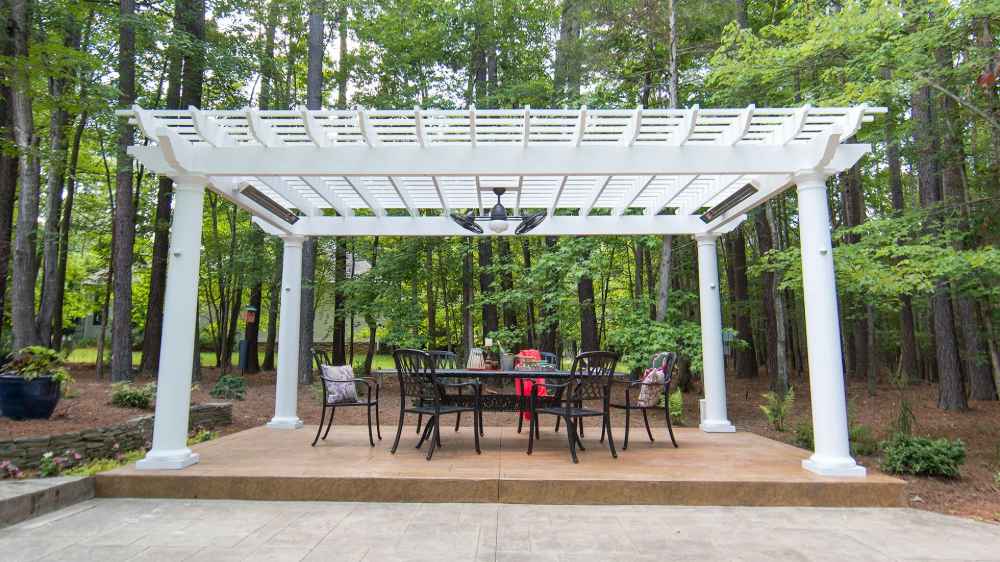 A centered view shows a black dining set under a white Structureworks Trex Pergola with built-in lighting and fans in a wooded area