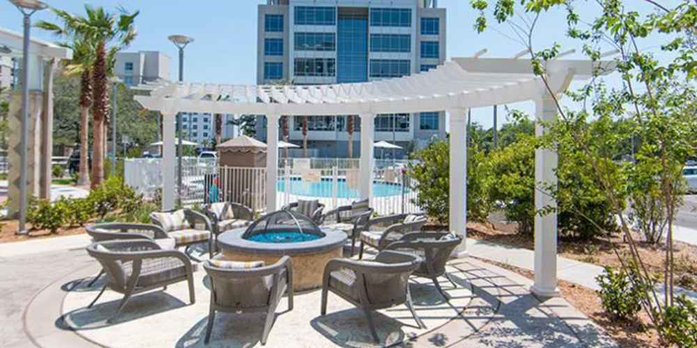 A circular fire pit is surrounded by cushioned chairs beneath a white Structureworks Trex Pergola near a pool in a sunny hotel courtyard