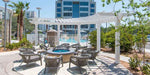 A circular fire pit is surrounded by cushioned chairs beneath a white Structureworks Trex Pergola near a pool in a sunny hotel courtyard