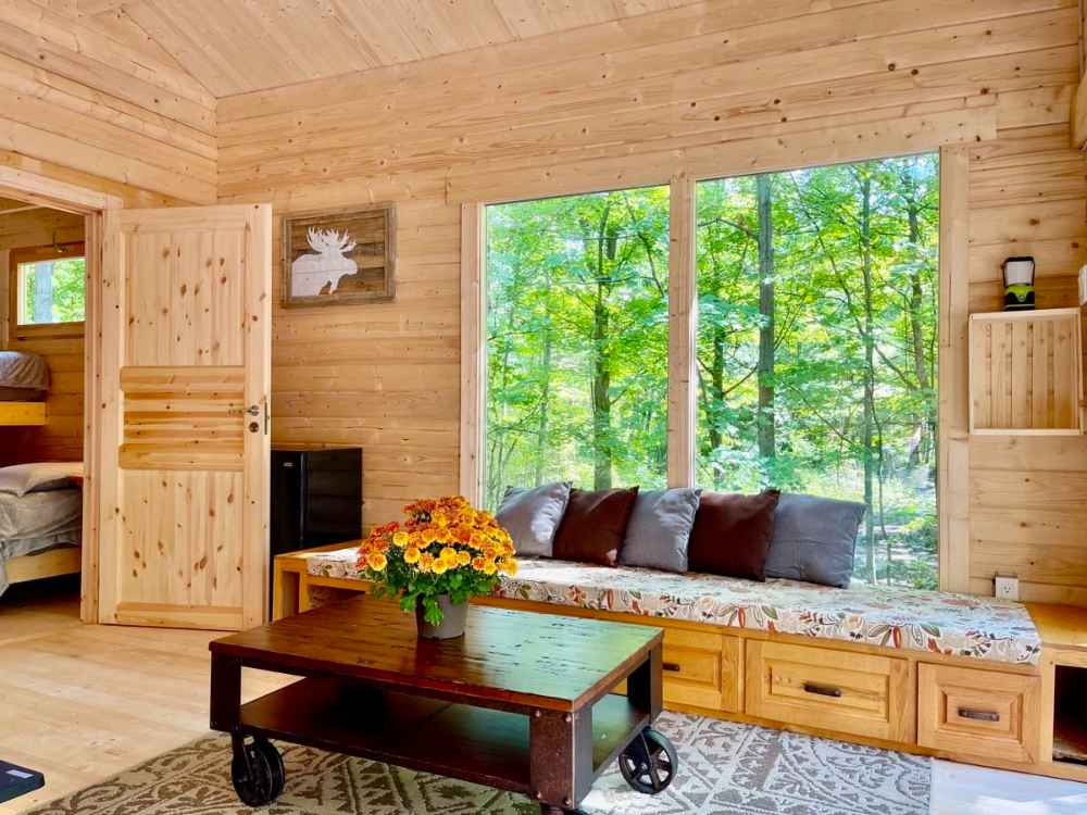 A cozy bench with storage and pillows faces large forest-view windows in the Sawmill Structures Edwin Lake House 70MM Garden Room