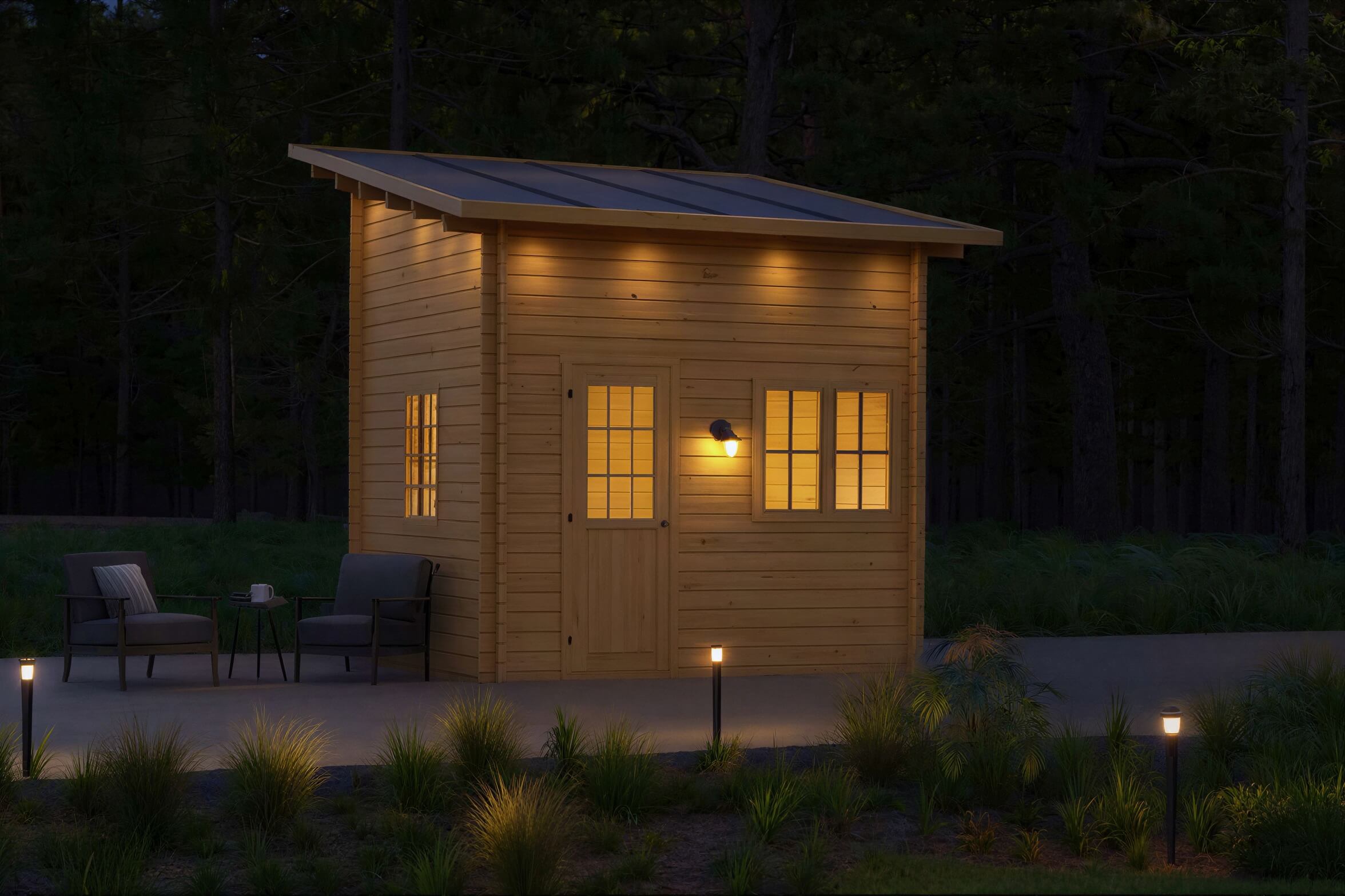 A cozy, illuminated Sawmill Structures Cube Loft 100 Garden Room glows warmly at night, surrounded by forest and outdoor seating