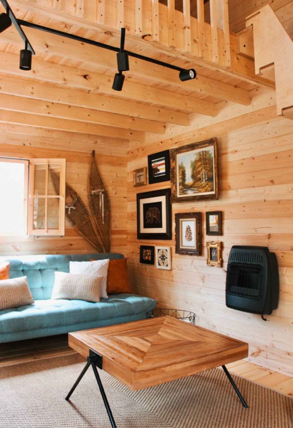 A cozy living space in the Sawmill Structures BlueWater Loft 160 70mm Garden Room includes a teal couch, rustic decor, and a geometric wood coffee table beneath loft stairs