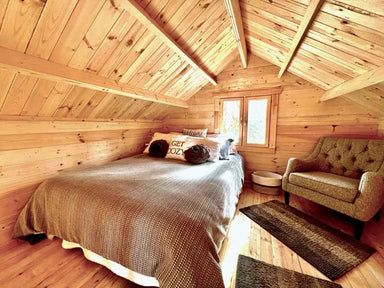 A cozy loft bedroom with a sloped wooden ceiling, soft lighting, and a green armchair is part of the Sawmill Structures The Jumbo Grand Loft 130 SD Garden Room