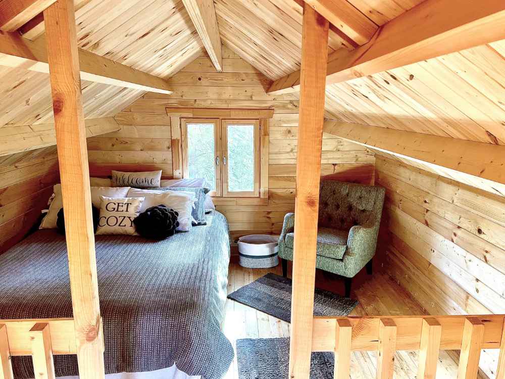 A cozy loft bedroom with sloped wooden ceilings, a full-size bed, accent pillows, and a green armchair is tucked inside the Sawmill Structures The Jumbo Grand Loft 130 SD Garden Room