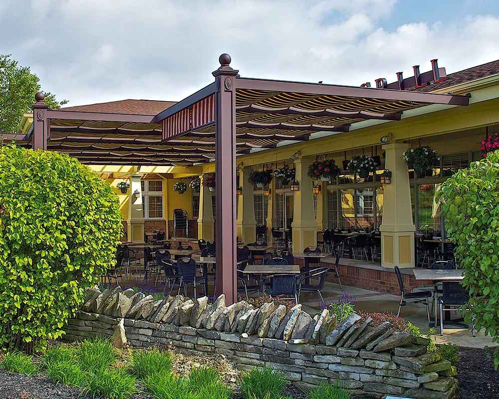 A cozy restaurant patio shaded by a brown-framed Structureworks Windsor Aluminum Patio Cover with striped fabric panels