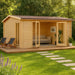A cozy wooden cabin with a sofa and plants inside, the GardenHouse24 USA Davos 44B Garden Room sits on a deck surrounded by lush green grass and trees
