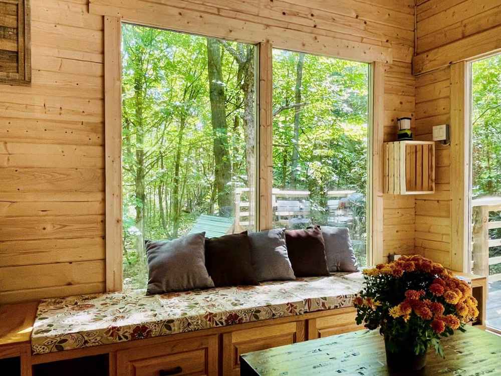 A cozy wooden window seat with floral cushions and gray pillows overlooks a lush green forest through large windows in the Sawmill Structures Edwin Lake House PLUS 70MM Garden Room