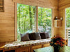 A cozy wooden window seat with floral cushions and gray pillows overlooks a lush green forest through large windows in the Sawmill Structures Edwin Lake House PLUS 70MM Garden Room