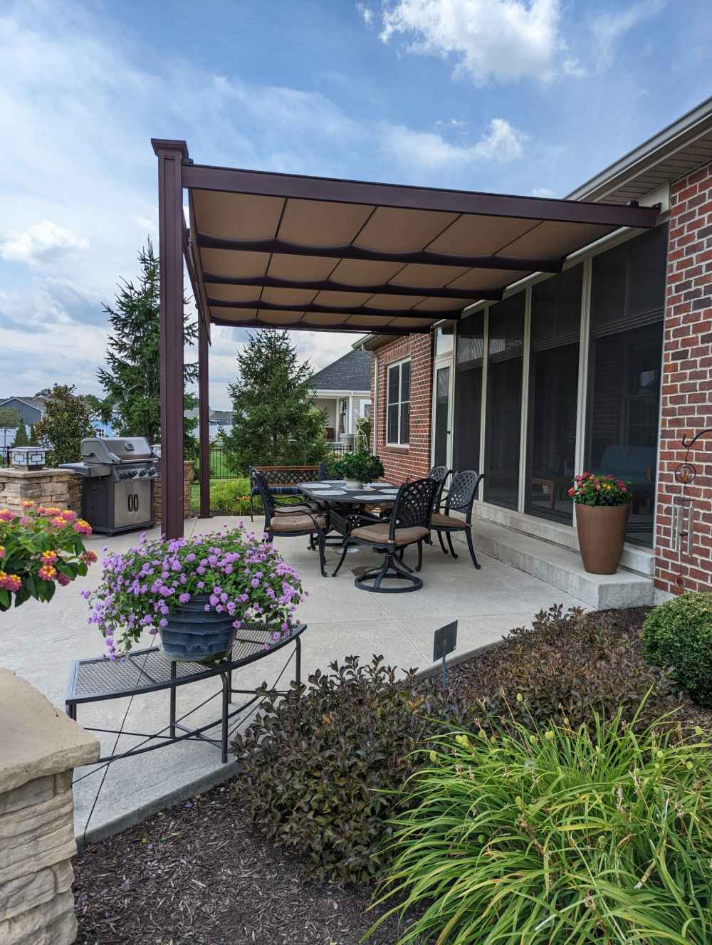 A flower-lined backyard dining space is covered by a modern brown-framed Structureworks Craftsman Aluminum Patio Cover & Pergola extending from a brick house