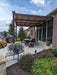 A flower-lined backyard dining space is covered by a modern brown-framed Structureworks Craftsman Aluminum Patio Cover & Pergola extending from a brick house
