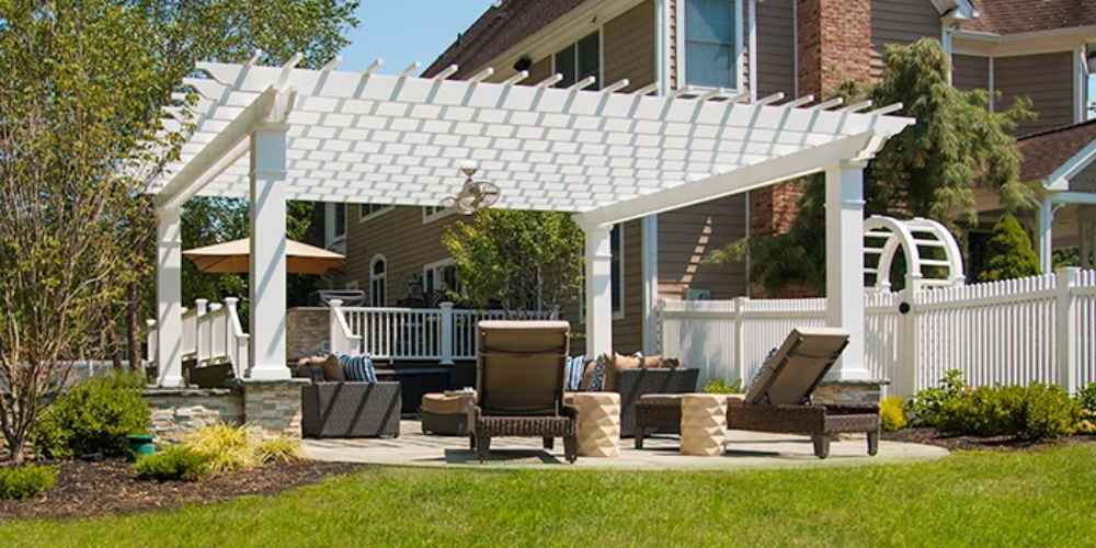 A garden seating area is shaded by a white Structureworks Williamsburg Fiberglass Pergola with fans and elegant columns over a stone base