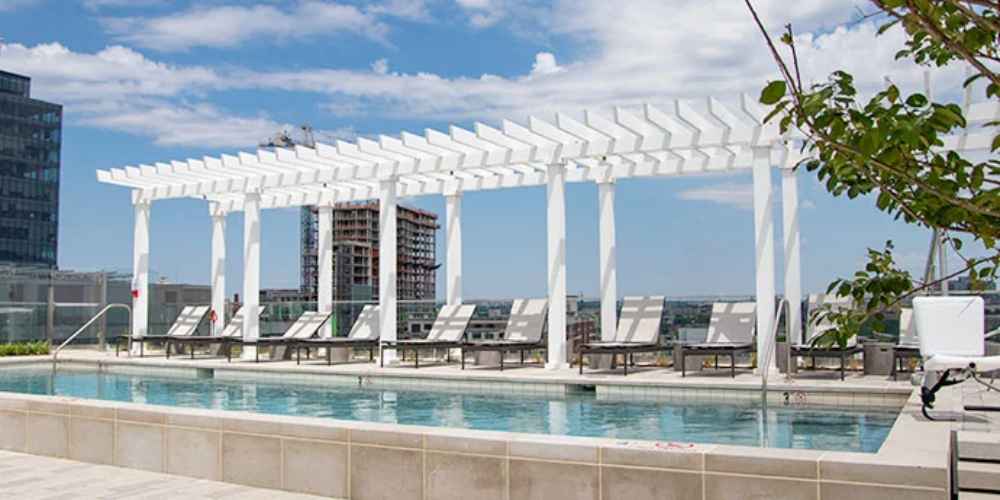 A long row of white posts supports a sleek Structureworks Williamsburg Fiberglass Pergola shading a rooftop pool deck with loungers beneath