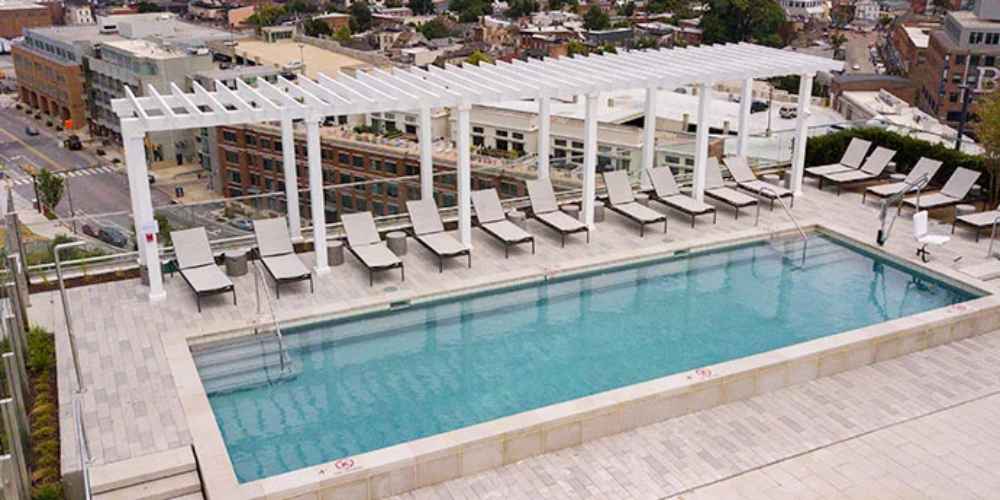 A minimalist Structureworks Williamsburg Fiberglass Pergola lines a rooftop terrace, casting shadows over poolside seating with a city view