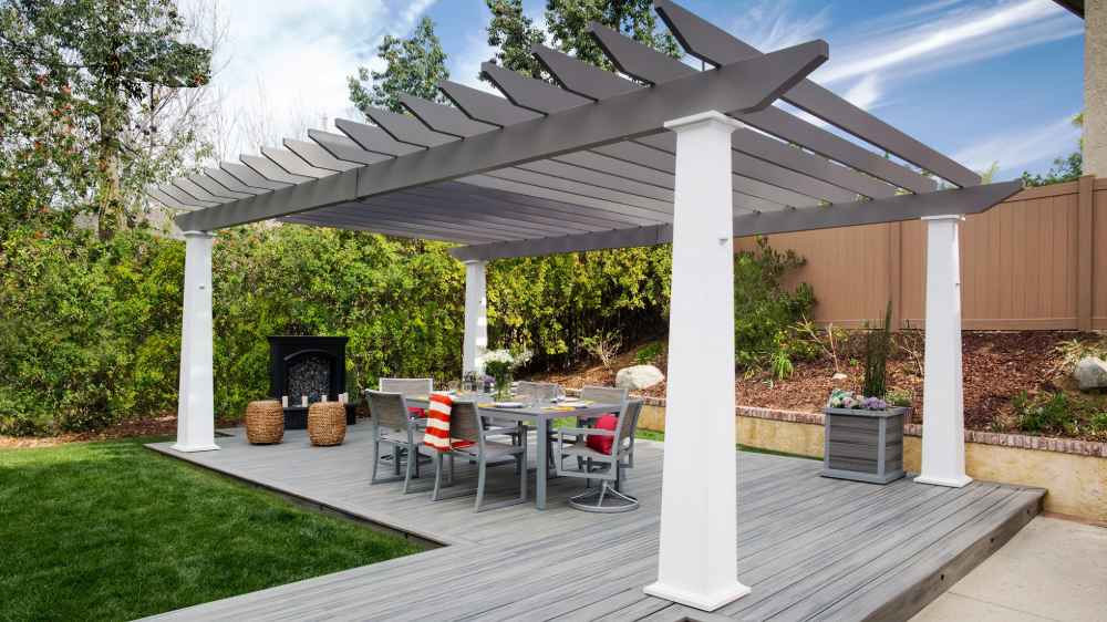 A modern dining setup with gray chairs rests on a wooden deck under a two-tone Structureworks Trex Pergola in a lush backyard