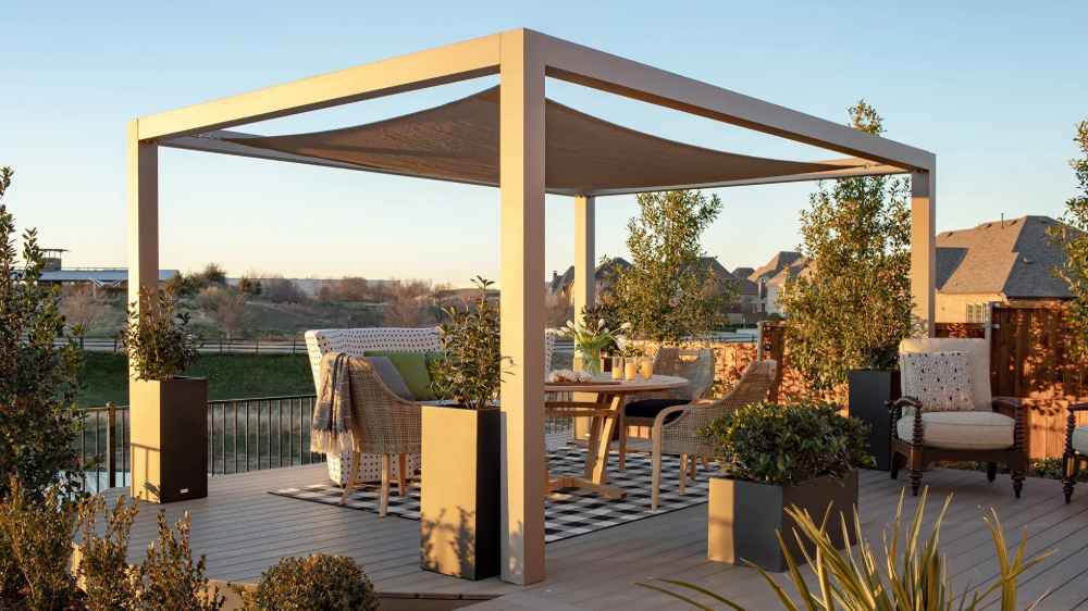 A modern outdoor patio features a beige Structureworks Solis Trex Pergola shading a cozy seating and dining area at sunset