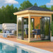 A modern wooden-framed glass GardenHouse24 USA Glaspavillon ALU 44D ISO Garden Room stands beside a pool with elegant furniture inside