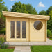 A modern wooden GardenHouse24 USA Sauna Tanonka 70 Garden Room with a circular side window and full glass double doors stands in a garden