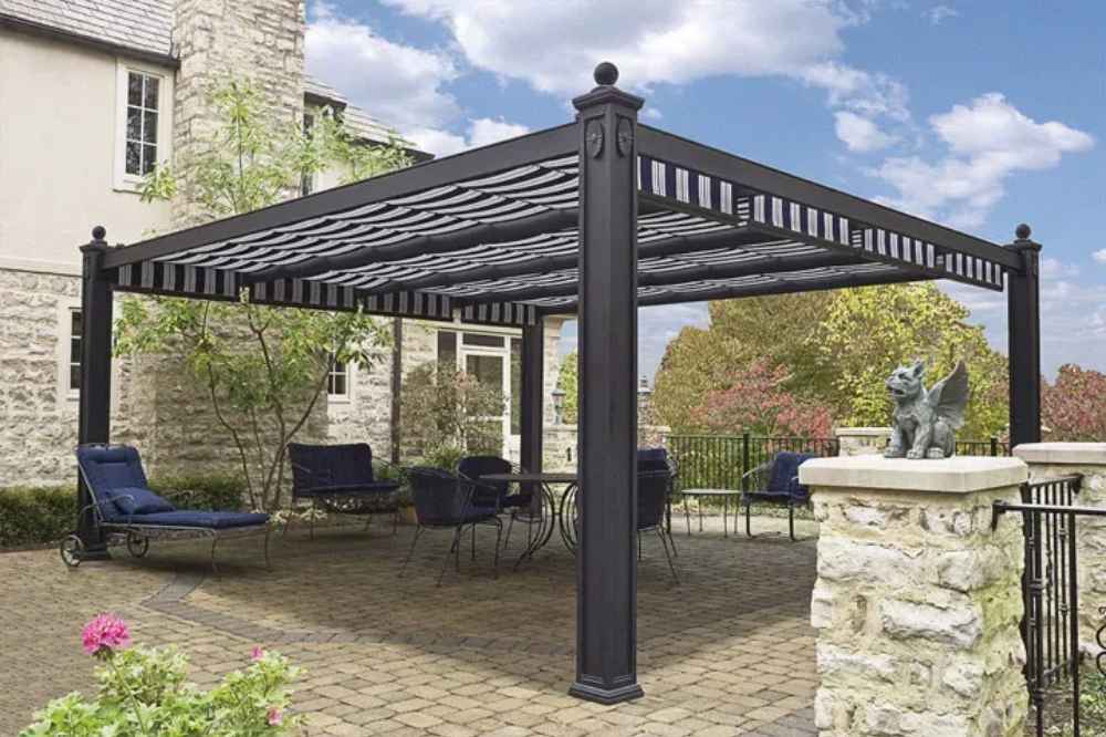 A paved courtyard beside a stone house furnished with dark blue patio furniture beneath a striped Structureworks Windsor Aluminum Patio Cover