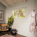 A peaceful corner of Sawmill Structures The Versa 240 70MM Garden Room displays a window with a forest view, a woven chair, and a hanging bathrobe