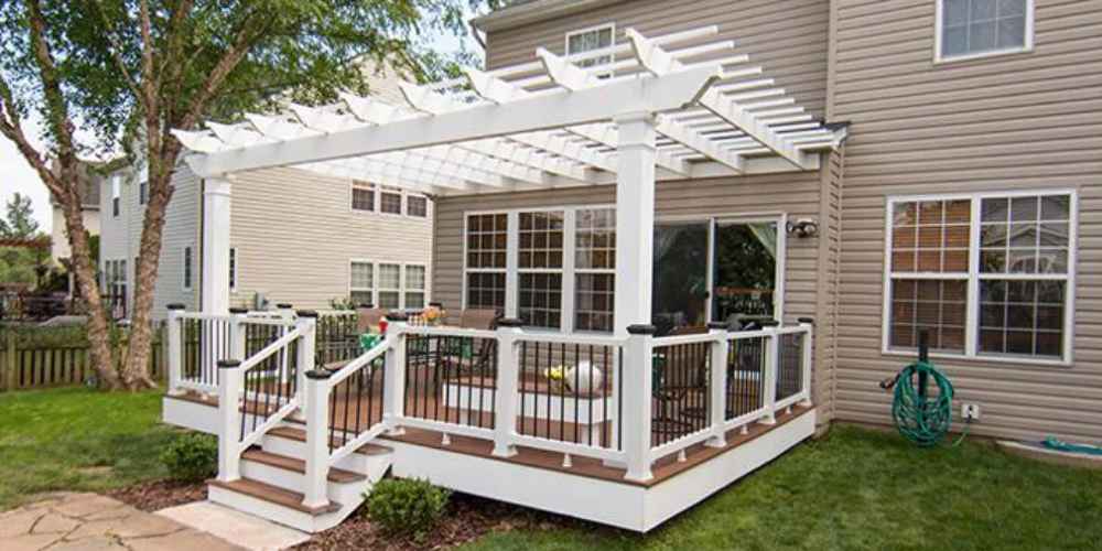 A raised backyard deck connects to a home and is covered by a classic white Structureworks Trex Pergola with railing