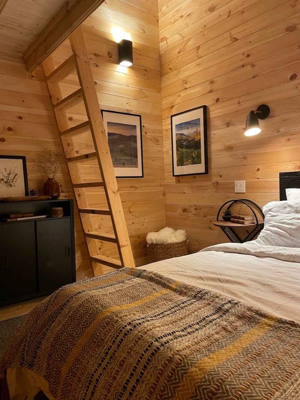 A rustic bedroom with a wooden ladder leading to a loft and warm-toned blankets showcases the inviting interior of the Sawmill Structures The Jumbo Grand Loft 130 SD Garden Room