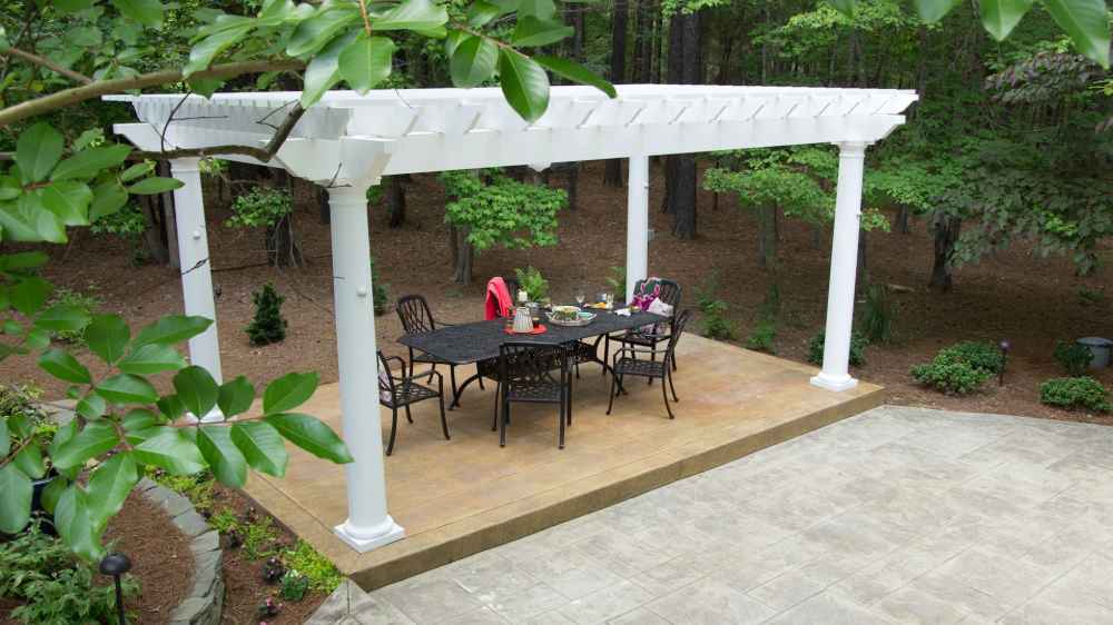 A secluded forest patio features a black dining table and chairs shaded by a white Structureworks Trex Pergola