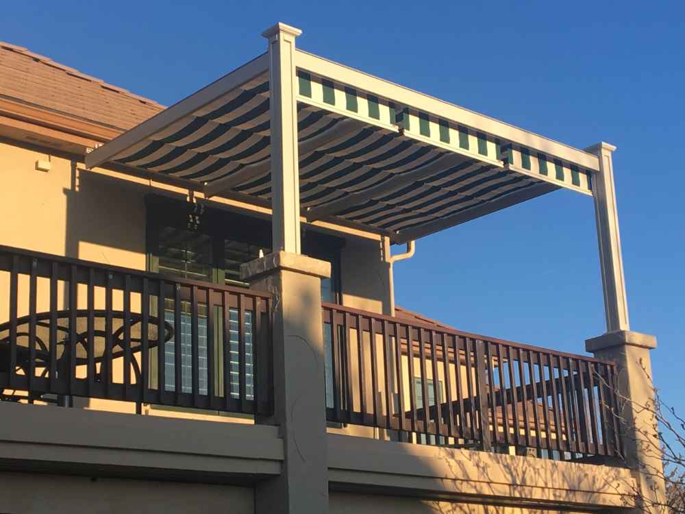 A second-story balcony is shaded by a striped retractable awning within a sleek, white-framed Structureworks Craftsman Aluminum Patio Cover & Pergola