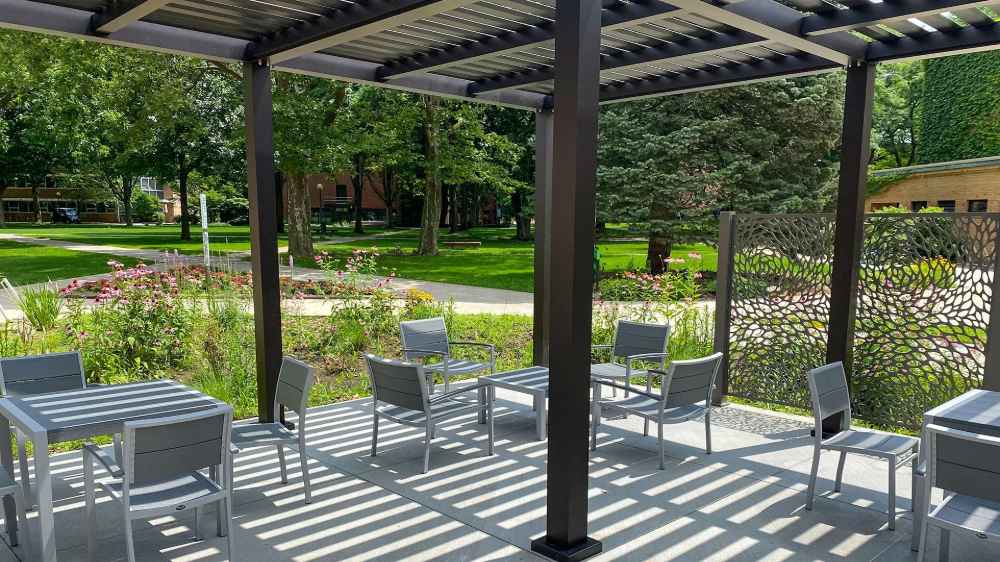 A shaded patio with modern gray furniture beneath a black Structureworks Element Trex Pergola offers a serene view of a flower garden and campus greenery