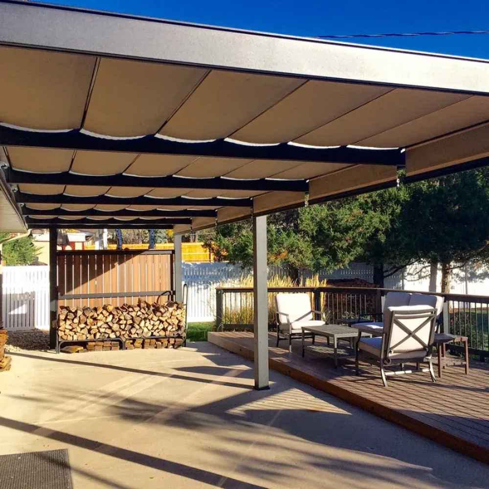 A silver Structureworks Outlook Aluminum Patio Cover & Pergola shades a concrete patio with stacked firewood and outdoor furniture beside a small deck