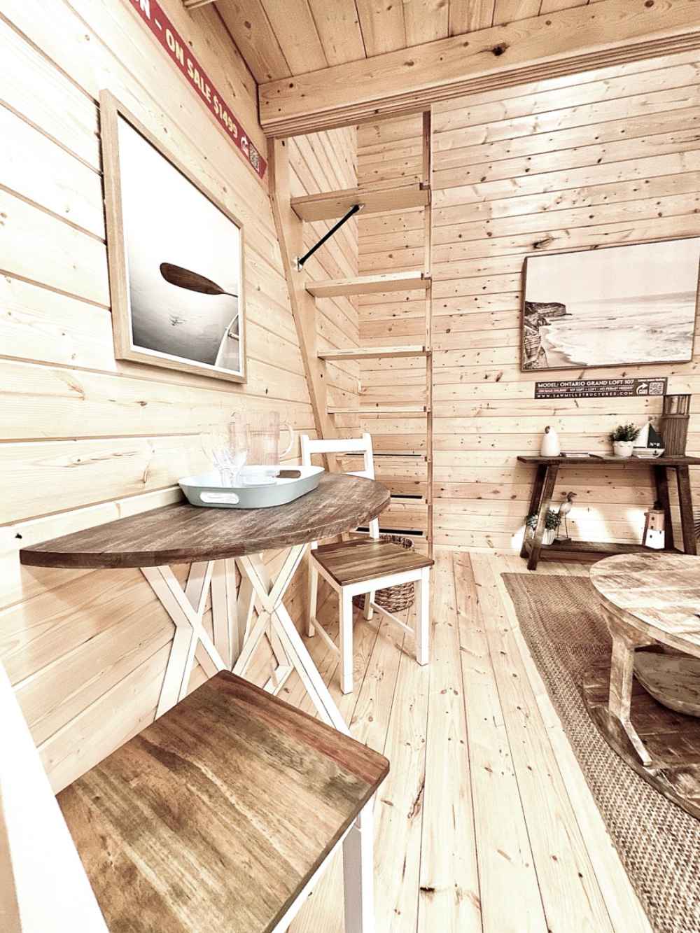 A small dining area with a round wood table and two stools sits beside a ladder staircase inside the Sawmill Structures Ontario Grand Loft 107 SD Garden Room
