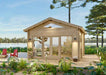 A small wooden cabin with glass doors and tall windows sits peacefully by a lake surrounded by greenery, part of the Sawmill Structures Edwin Lake House PLUS 70MM Garden Room